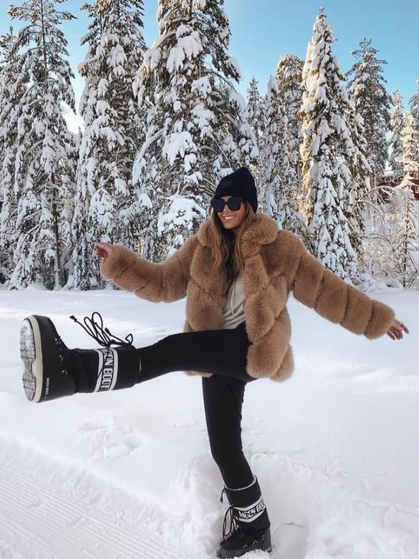 Woman in black Moon Boots, camel faux-fur coat and black leggings playfully kicking in snow