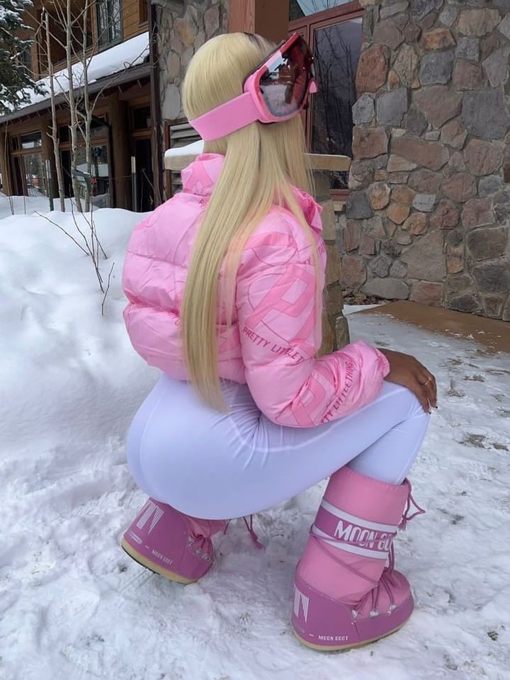 Woman in pink Moon Boots, pink puffer jacket, white leggings and ski goggles crouching in snow