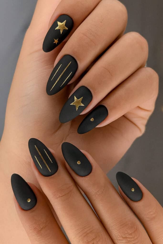 Matte black almond nails with gold foil stars, stripes, and dot accents