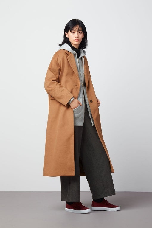 Woman in a camel coat over a grey hoodie with dark wide-leg trousers and red sneakers