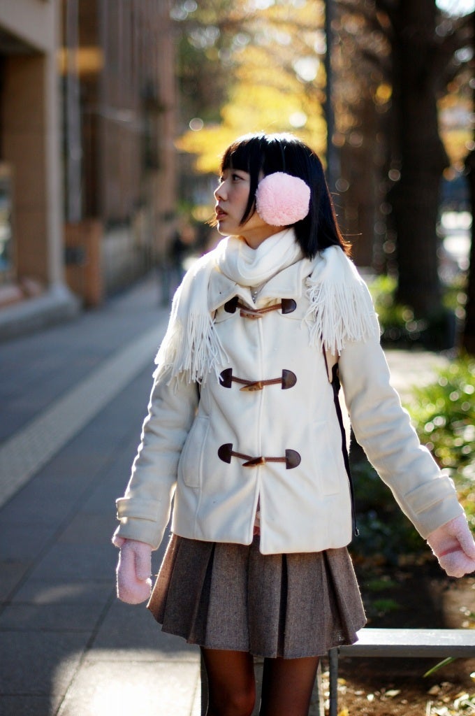 Woman wearing pink fluffy earmuffs, white toggle duffle coat, pleated skirt, and pink gloves on a city street