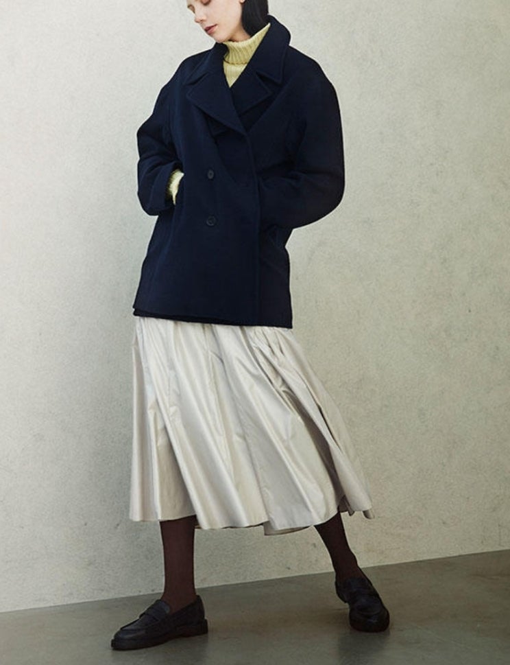 Woman in navy double-breasted peacoat, pleated white midi skirt, dark tights and black loafers