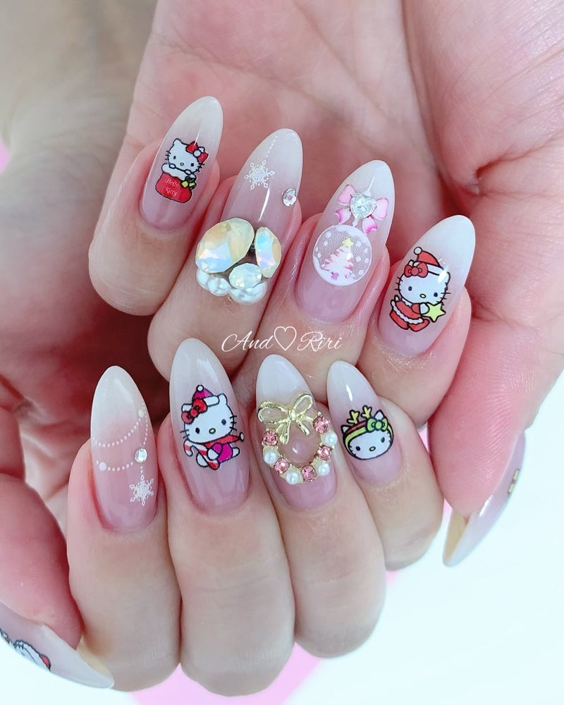 Medium almond nails in sheer nude with Hello Kitty in a Santa outfit, iridescent gemstone clusters, a pearl and gold bow wreath and snowflakes