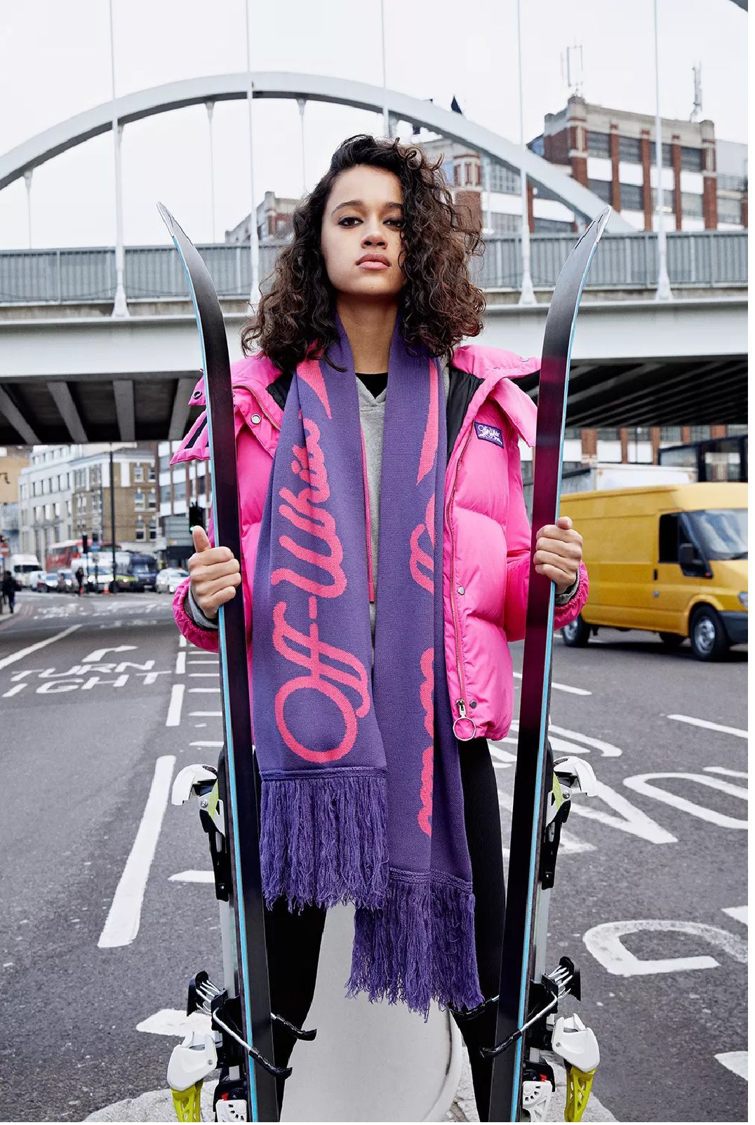 Woman holding skis in a city street, wearing a pink puffer jacket and a long purple logo scarf with fringe