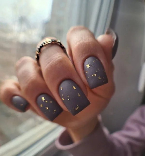 Short squoval nails in matte dark gray scattered with small gold foil flakes