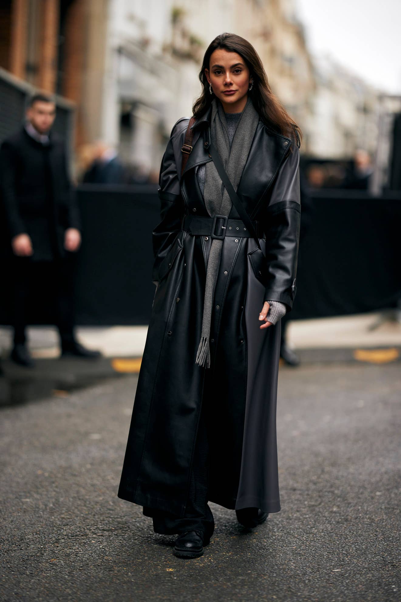 Woman in a black belted leather trench coat with a grey scarf and black boots at Paris Fashion Week