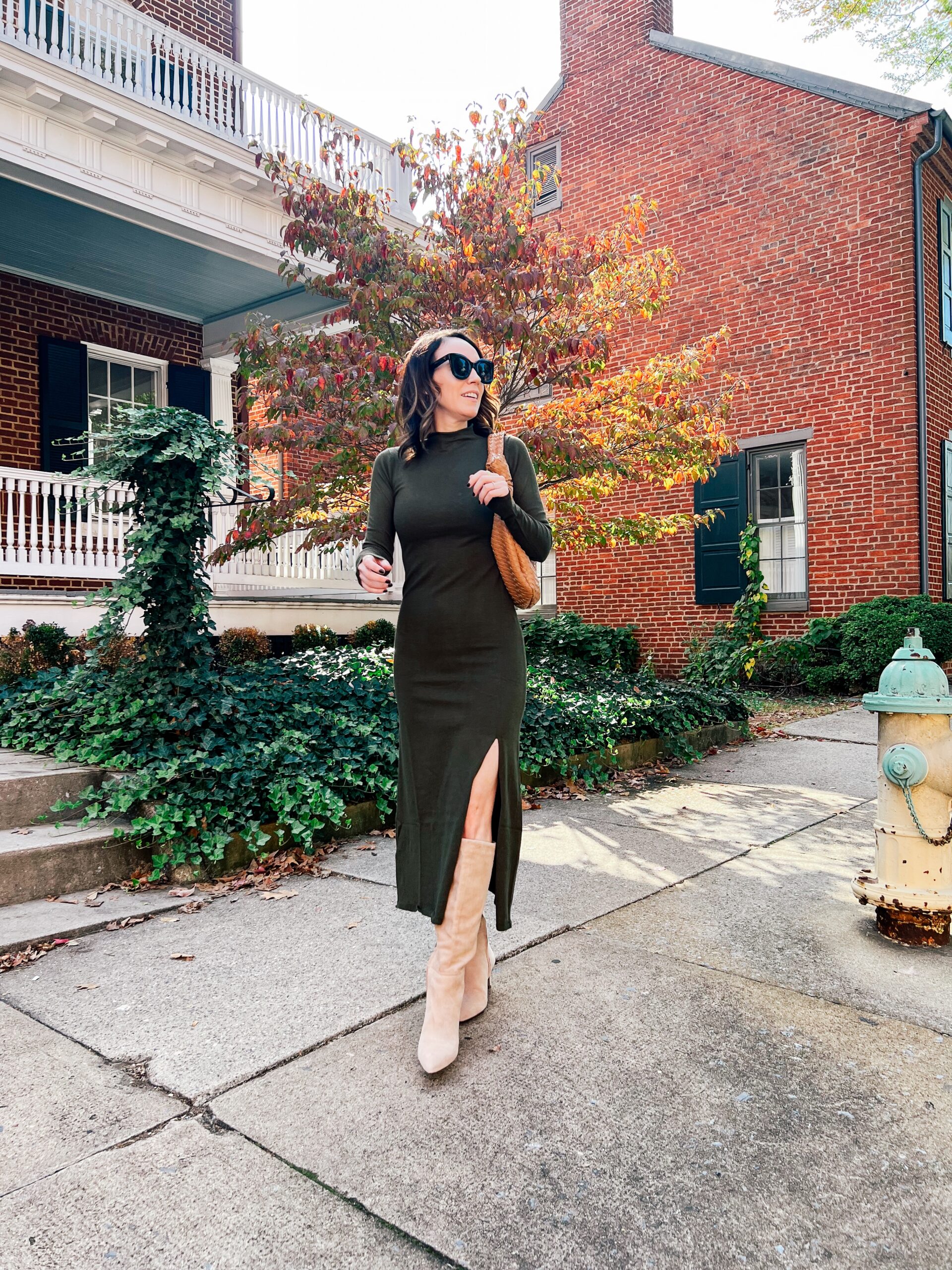 Dark olive mock-neck sweater dress with a side slit, tan suede knee boots, a woven shoulder bag and sunglasses