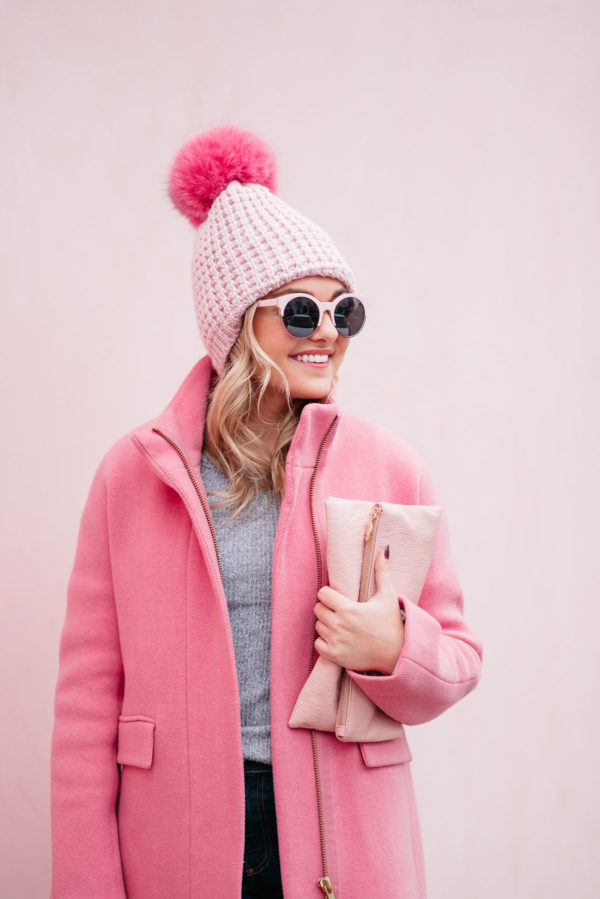 Woman in a pink pom beanie and pink coat with sunglasses and clutch