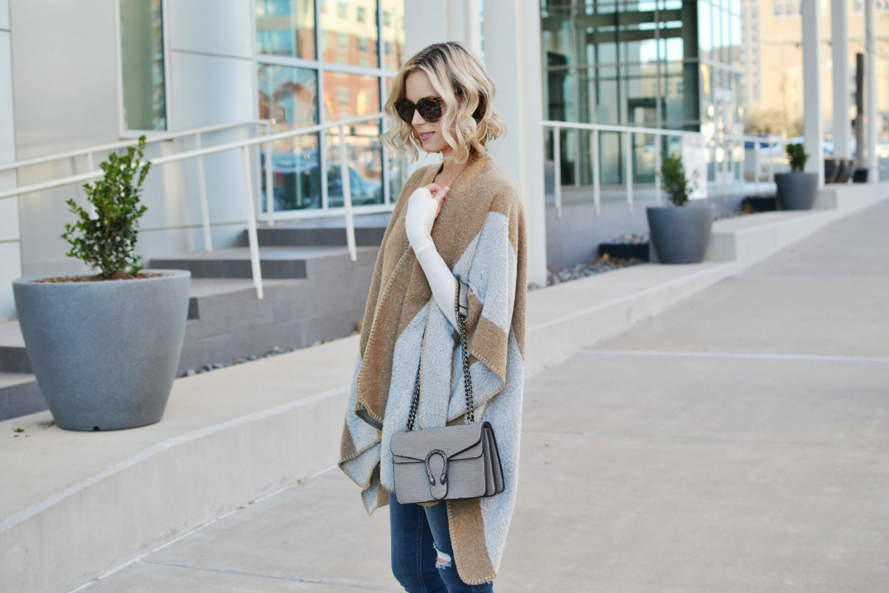 Woman in a tan and gray knit poncho over a white thermal with jeans and a gray handbag