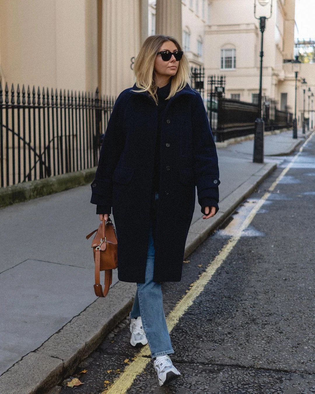 Woman in a navy wool coat with blue jeans, white sneakers and a tan leather bag on a city street