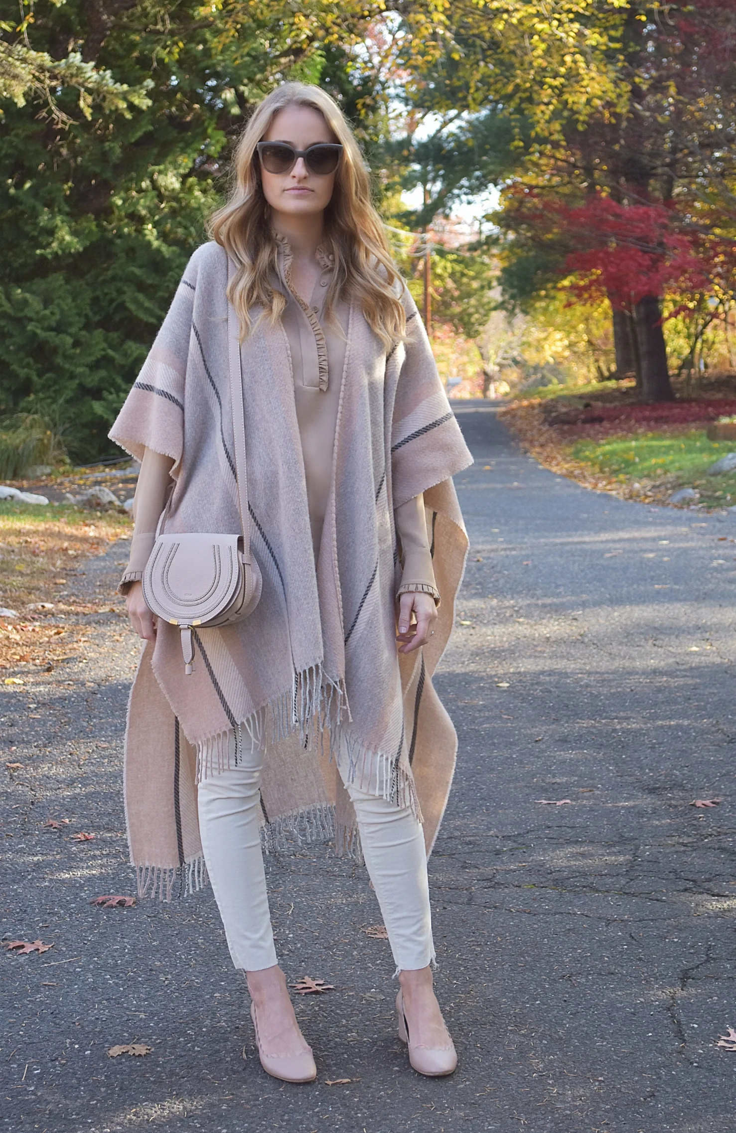 Woman in a beige striped fringed cape over a beige ruffled blouse, white jeans and nude pumps