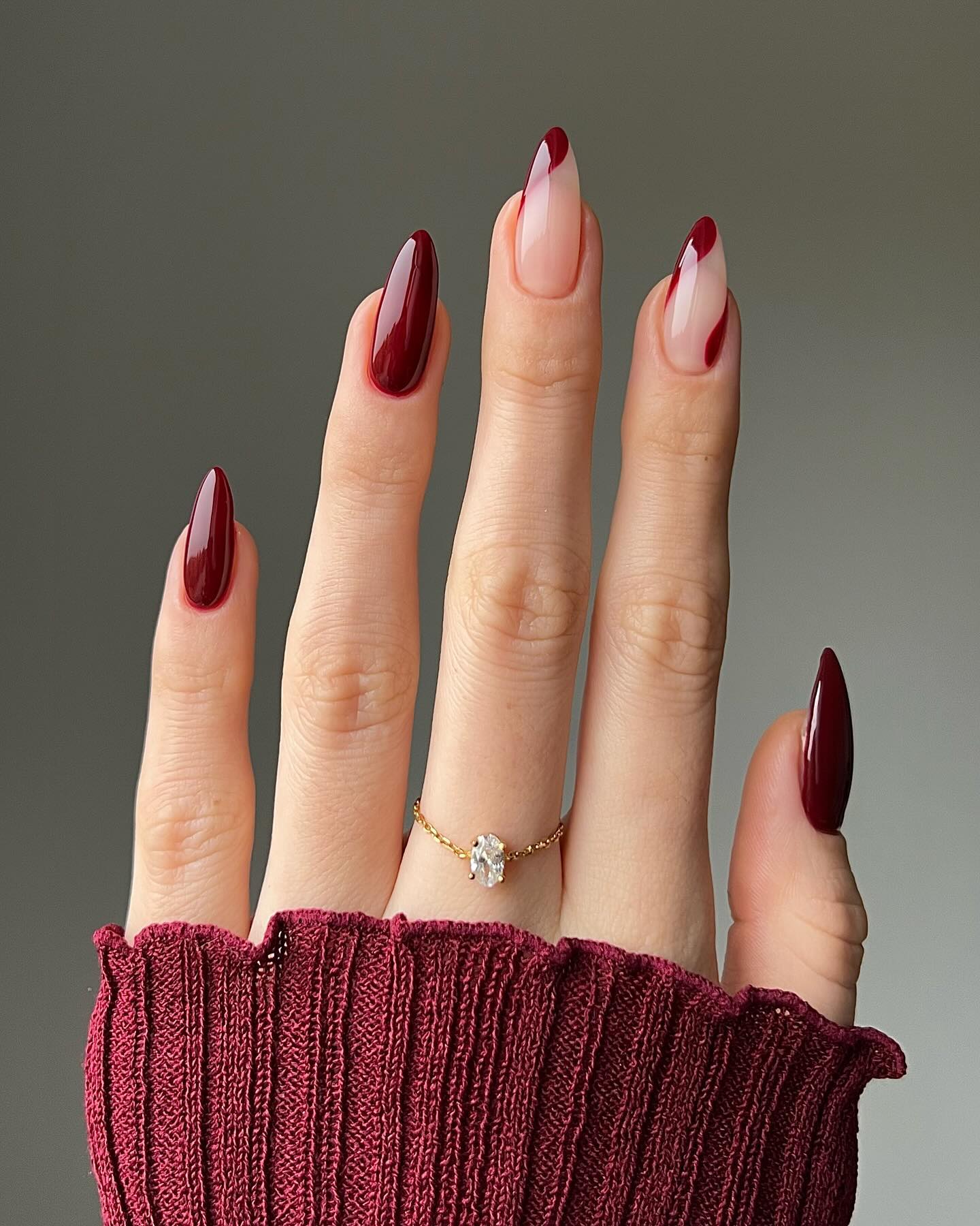 Long almond nails in deep wine red with two nude French-tip accent nails and a delicate gold ring