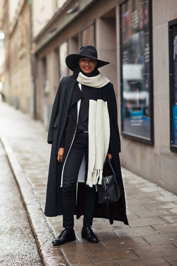 Woman wearing a black coat with a cream knit scarf, black wide-brim hat and black skinny pants