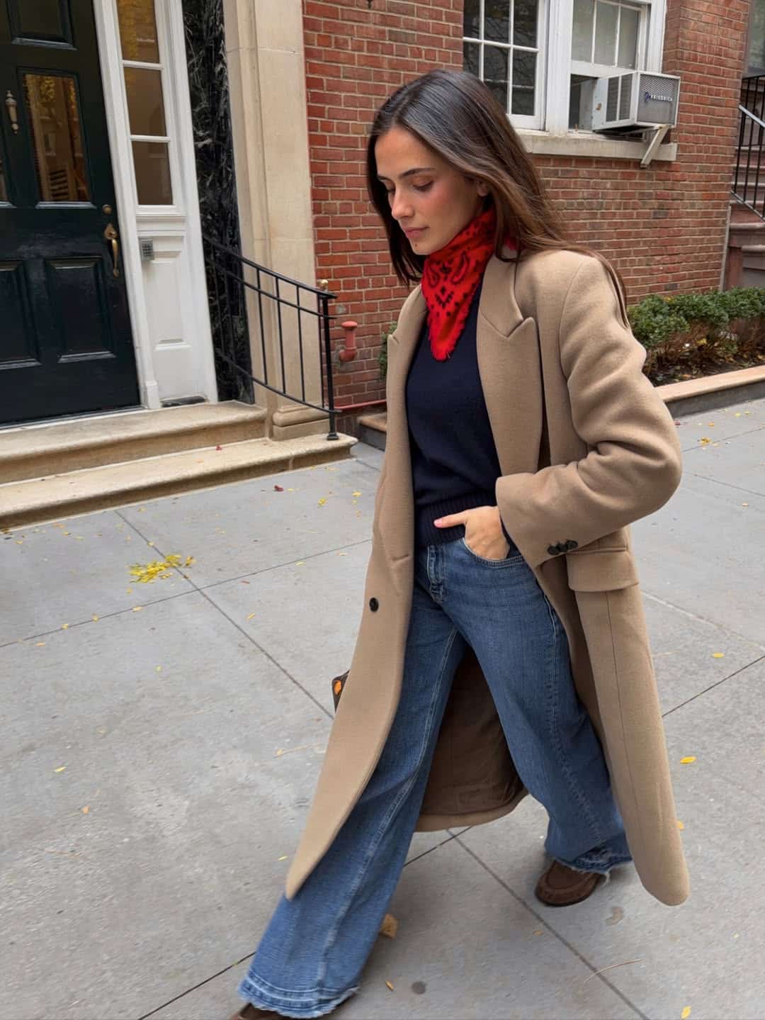 Woman in a camel coat with a red scarf and jeans in a winter street style look