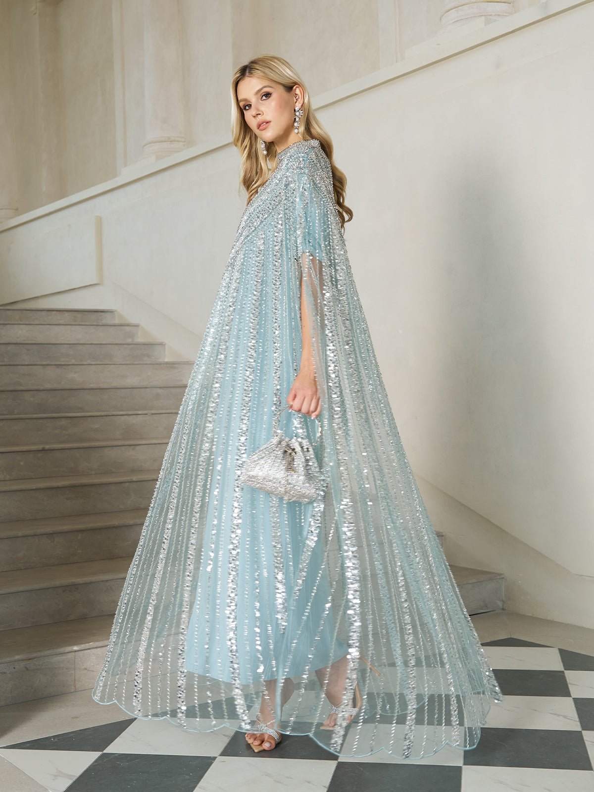 Woman wearing an ice-blue sequin cape gown with a silver mini bag and strappy heels