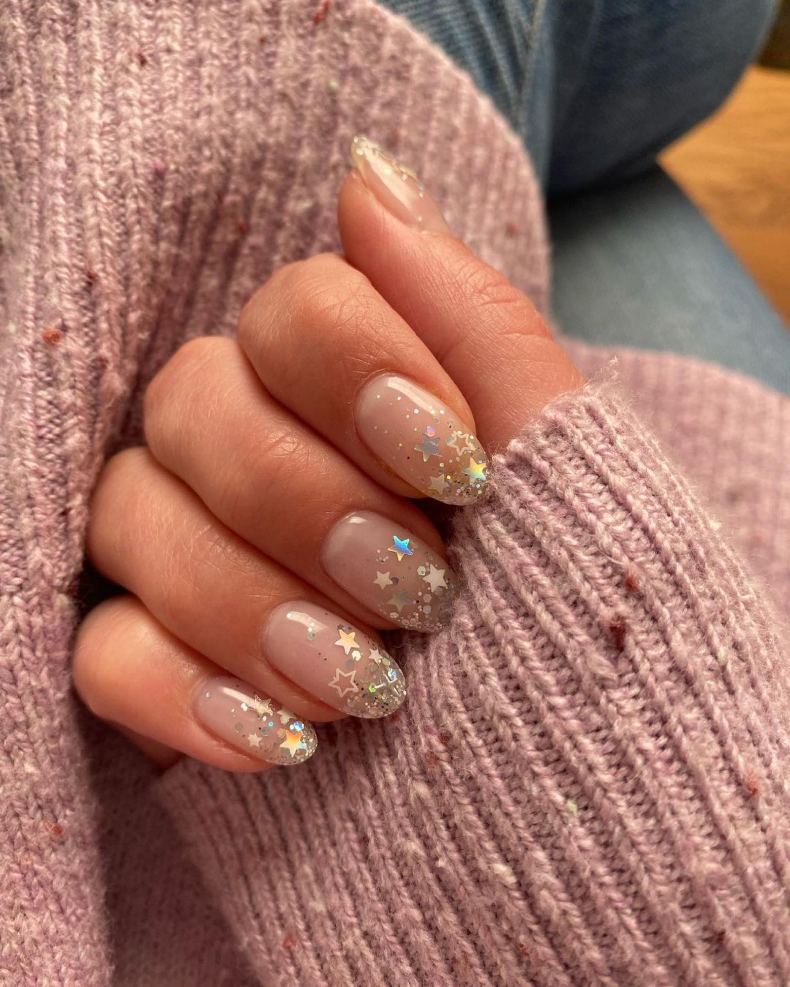 Sheer nude almond nails with silver glitter and holographic star confetti fading from the tips
