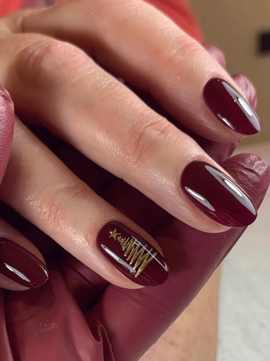 Glossy deep burgundy oval nails with a small gold Christmas tree drawn on one accent nail