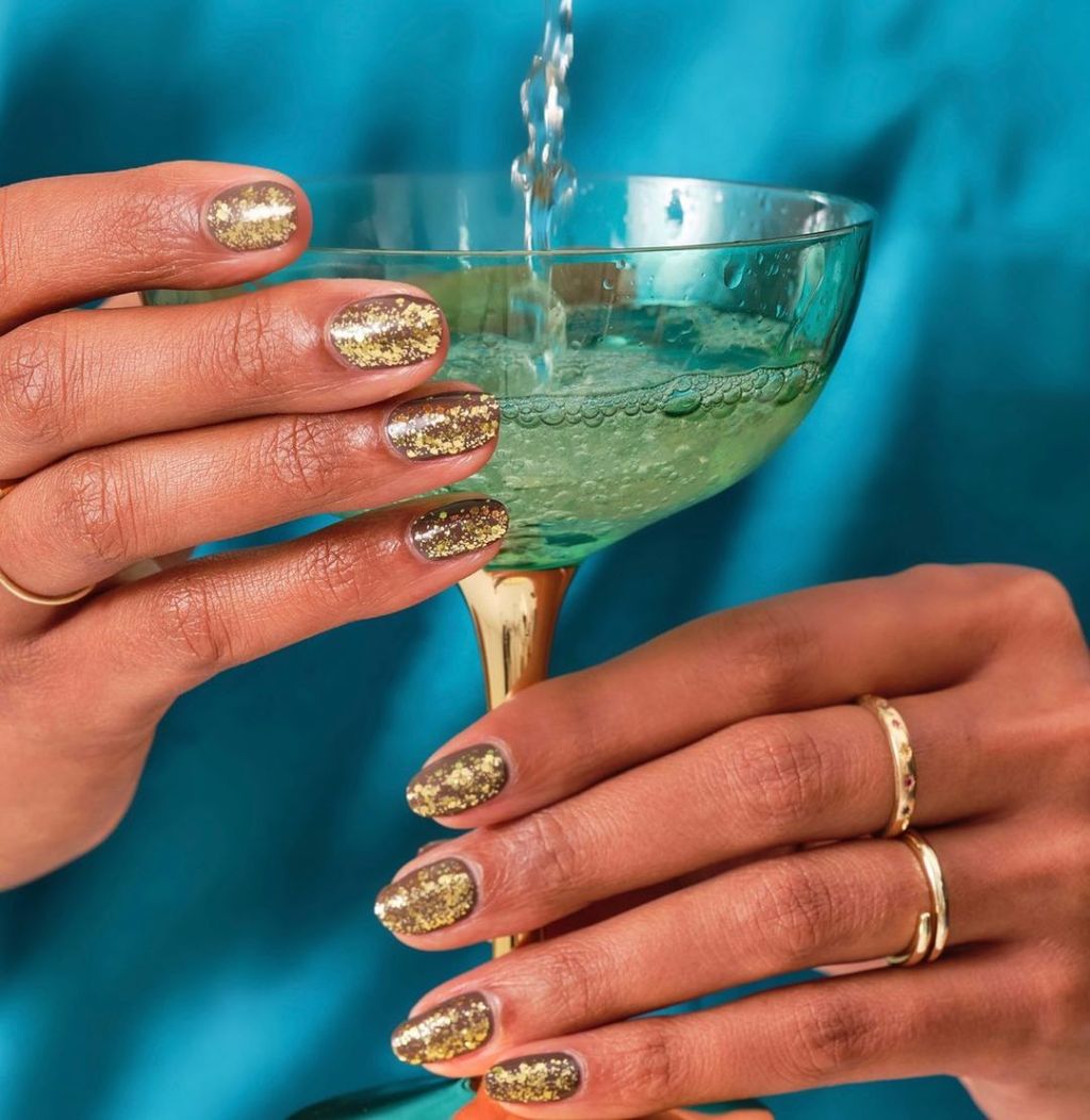 Short squoval nails in full-coverage gold glitter polish, hand holding a champagne coupe
