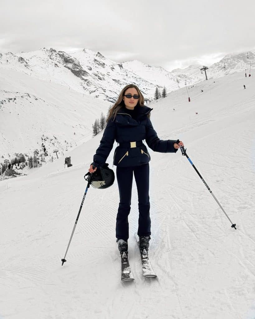 Woman standing on a ski slope in a navy belted puffer jacket and navy ski pants, holding a helmet and poles