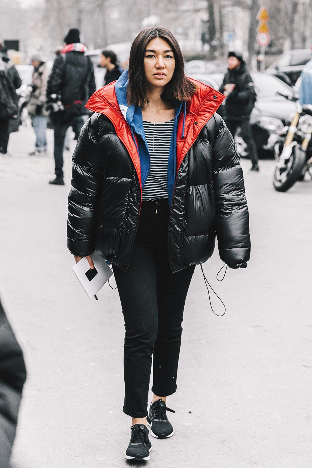 Woman in a glossy black puffer with a red and blue lined hood, striped tee, black jeans and black sneakers