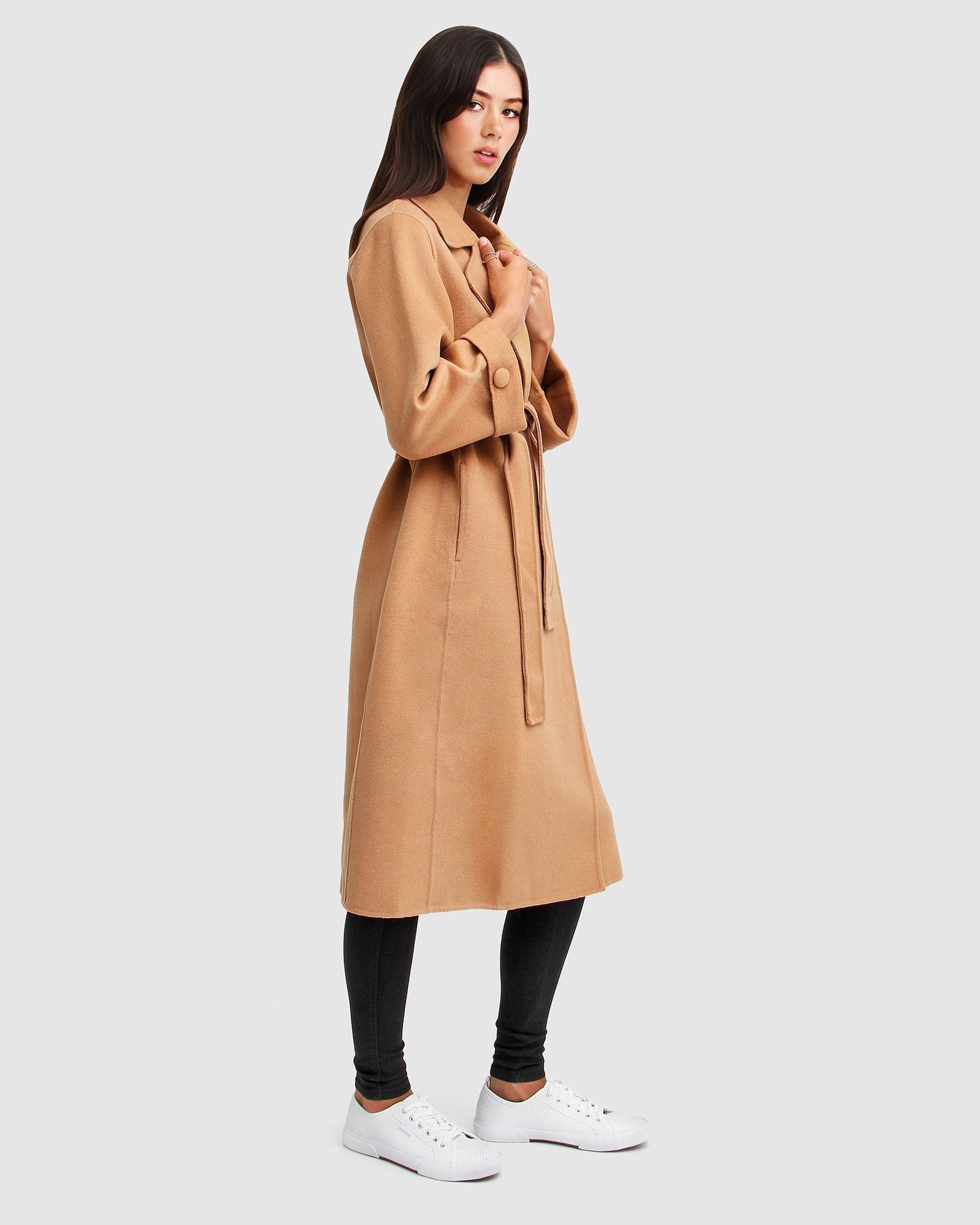 Model wearing a camel belted wool coat with black leggings and white sneakers