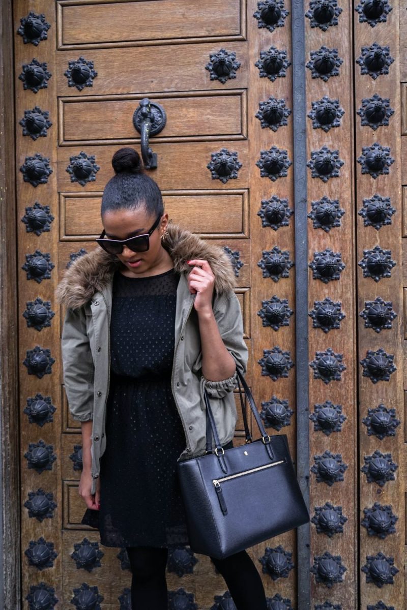 Woman in a sage fur-trim parka over a black dress carrying a large tote