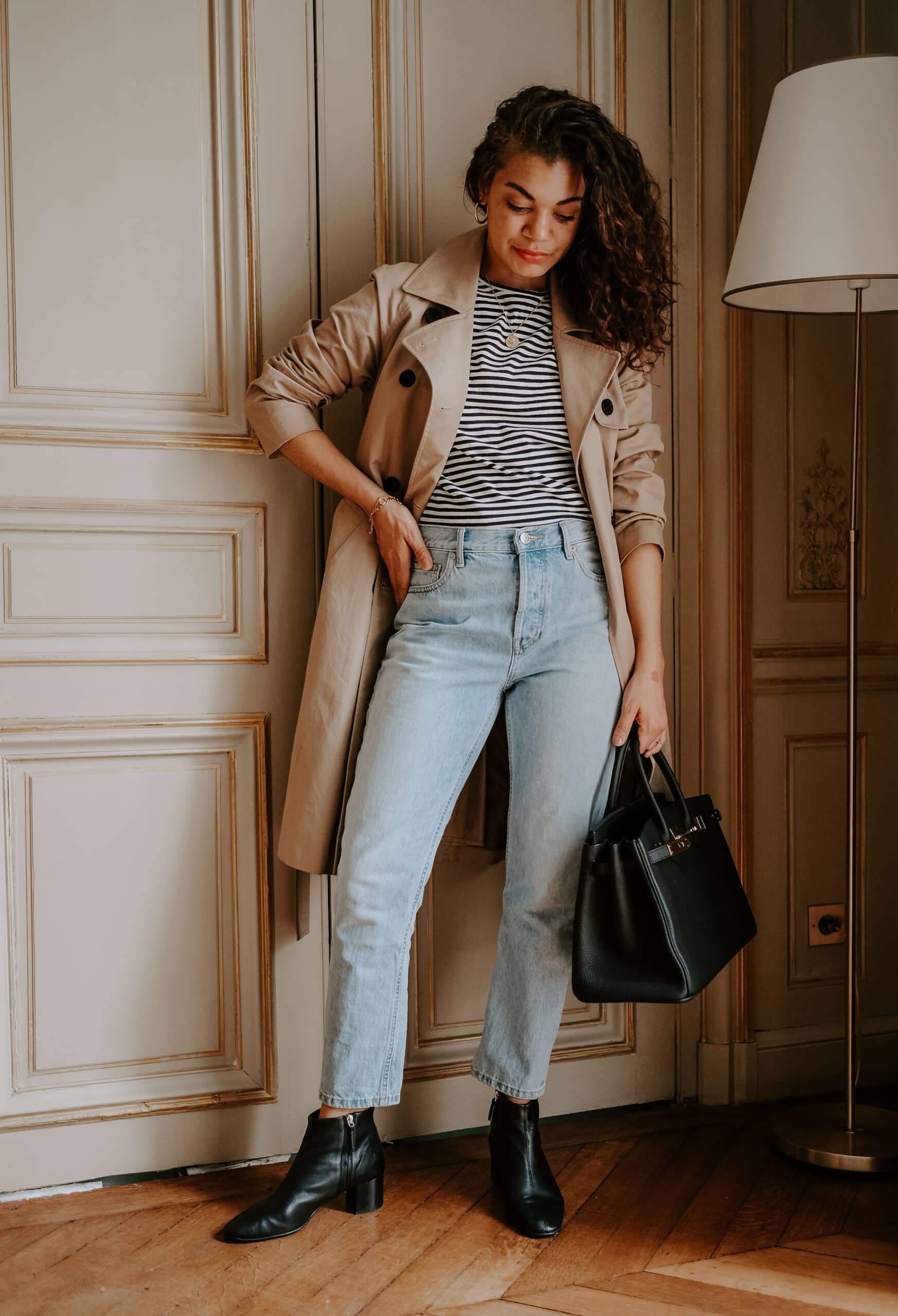 Woman in a beige trench coat over a navy and white striped tee with light-wash cropped jeans, black ankle boots and a black handbag