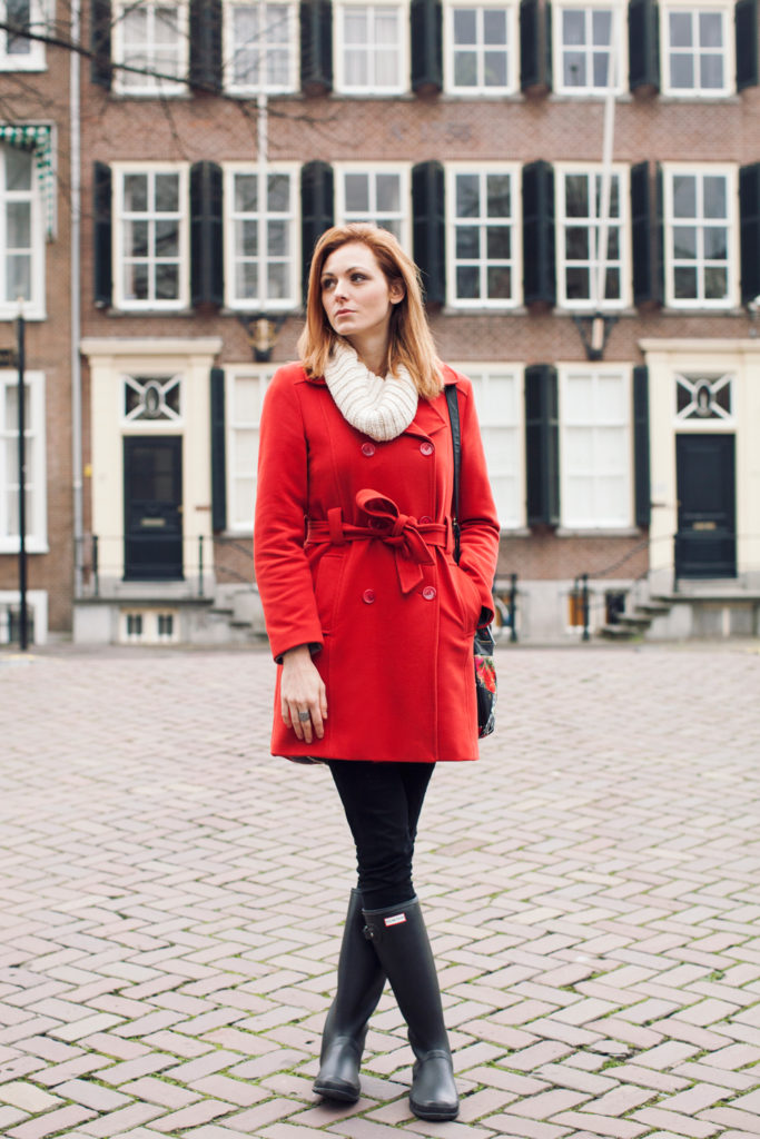 Woman in a red peacoat with white scarf, black skinny pants and black Hunter rain boots on a cobblestone street