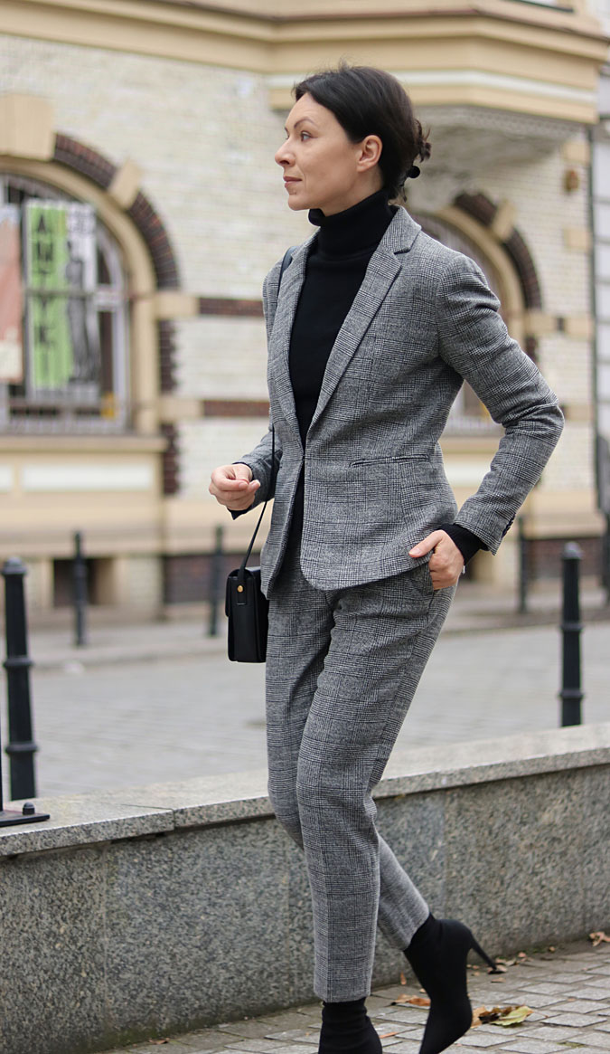 Woman on a city street wearing a grey houndstooth suit with a black turtleneck, black ankle boots and a small black bag