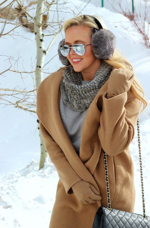 Woman wearing grey faux fur earmuffs, a camel wrap coat, and sunglasses in the snow