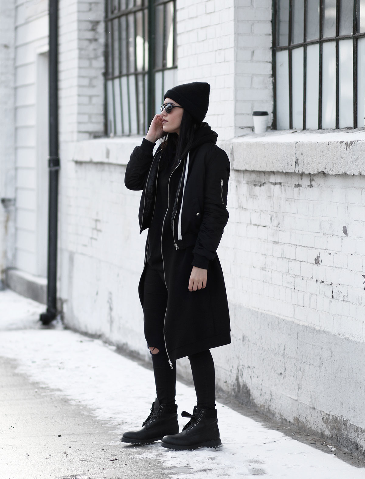 Woman in an all-black outfit with a bomber jacket, ripped skinny jeans, beanie, and black Timberland boots in the snow