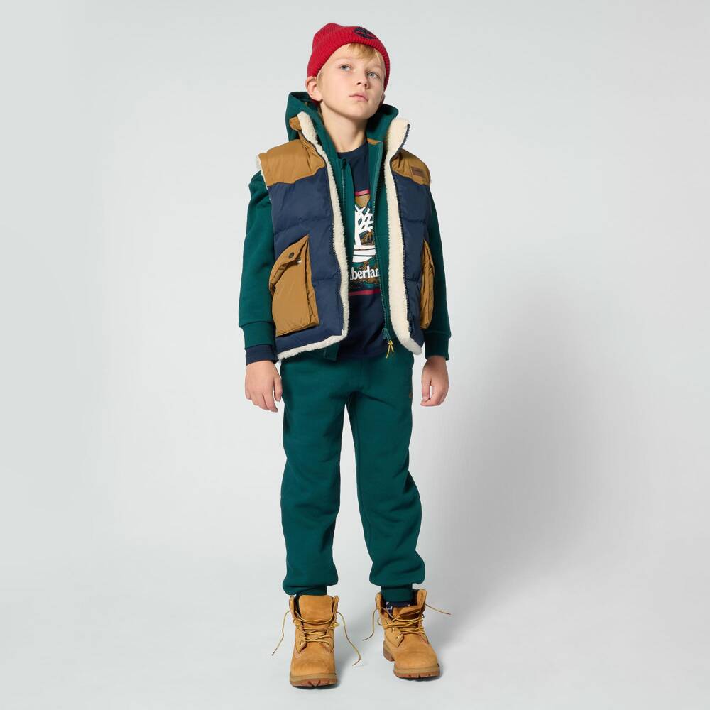 A boy wearing a sherpa-lined puffer vest over a green sweatshirt with joggers, a red beanie and tan boots