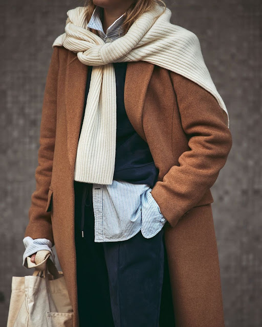 Woman wearing a cream ribbed knit scarf wrapped generously over a camel wool coat with a pinstripe shirt
