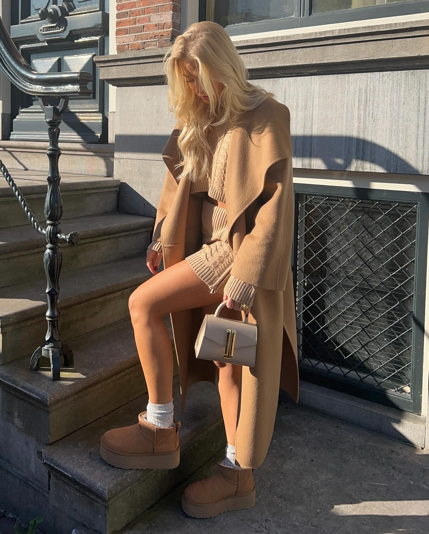 Woman wearing chestnut Ugg mini boots with a long camel coat over a ribbed knit midi dress