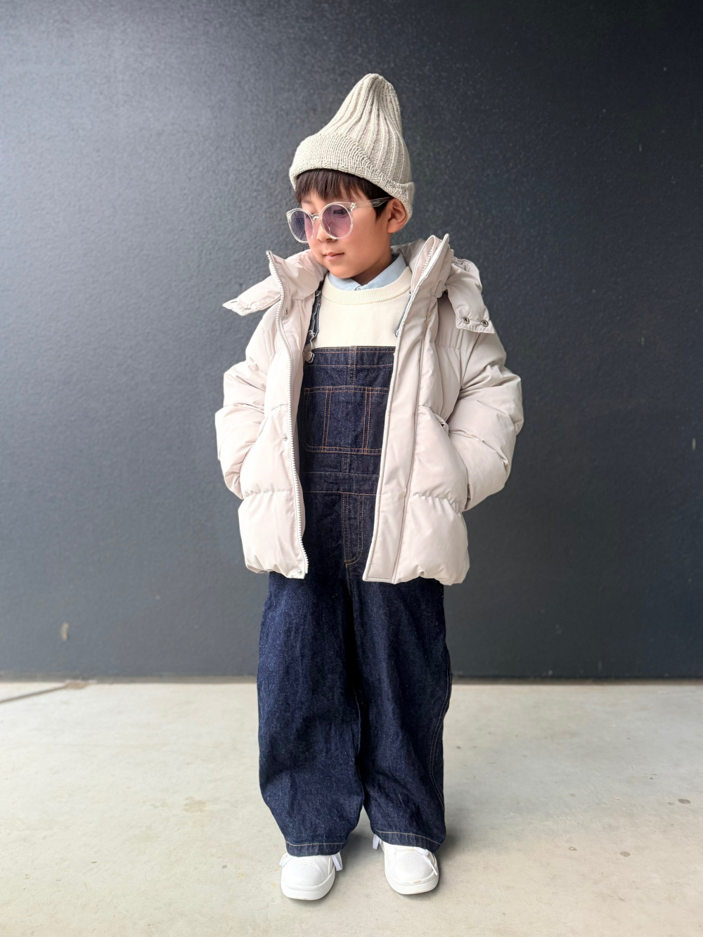 A child wearing a cream puffer jacket over dark denim overalls with a knit beanie and white sneakers