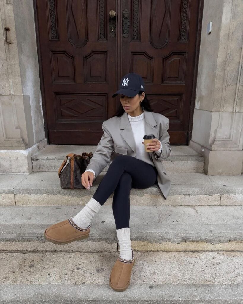 Woman in a grey blazer and black leggings wearing chestnut ugg tasman slippers with white socks and a baseball cap