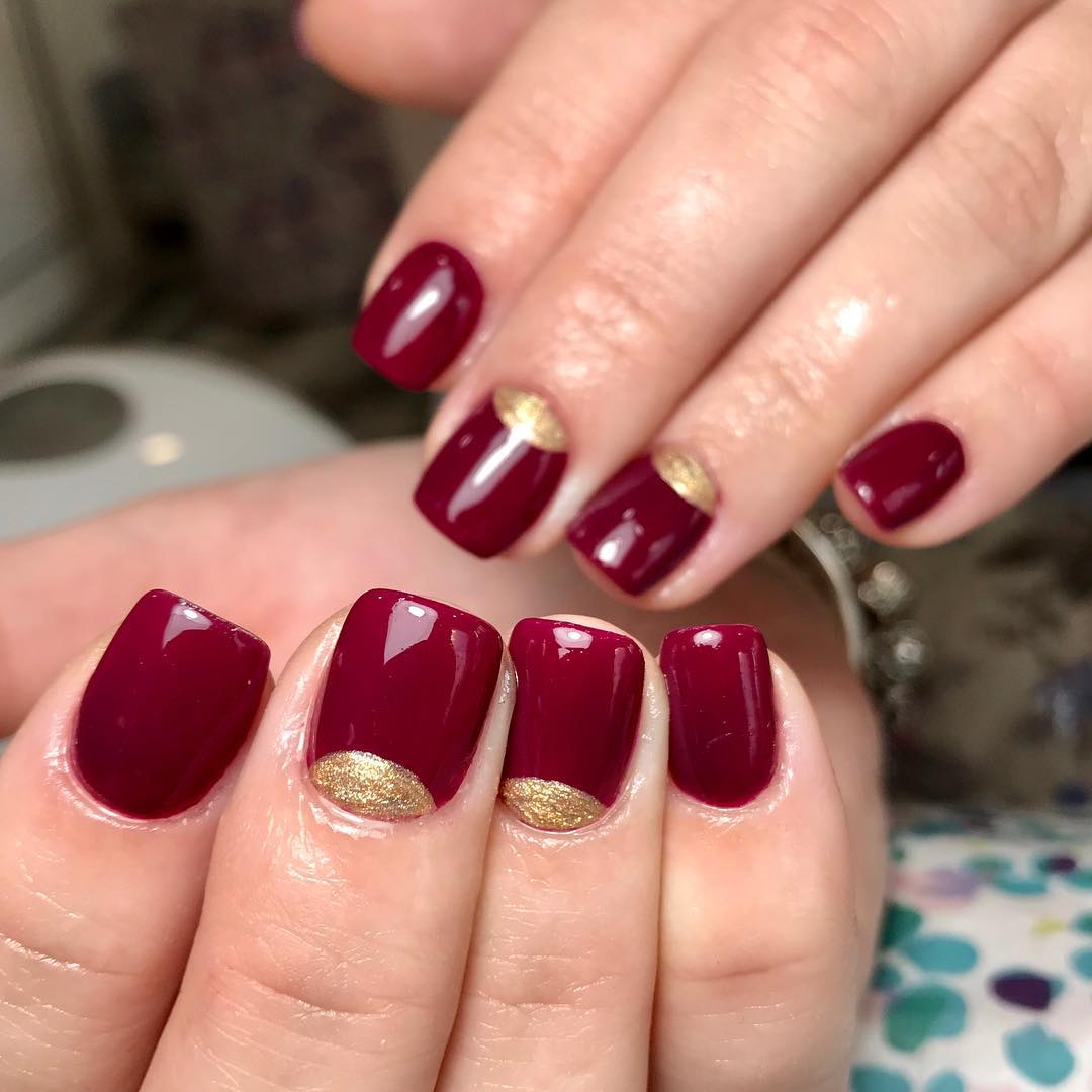 Short square burgundy nails with gold glitter half-moon accents at the cuticle on alternating nails