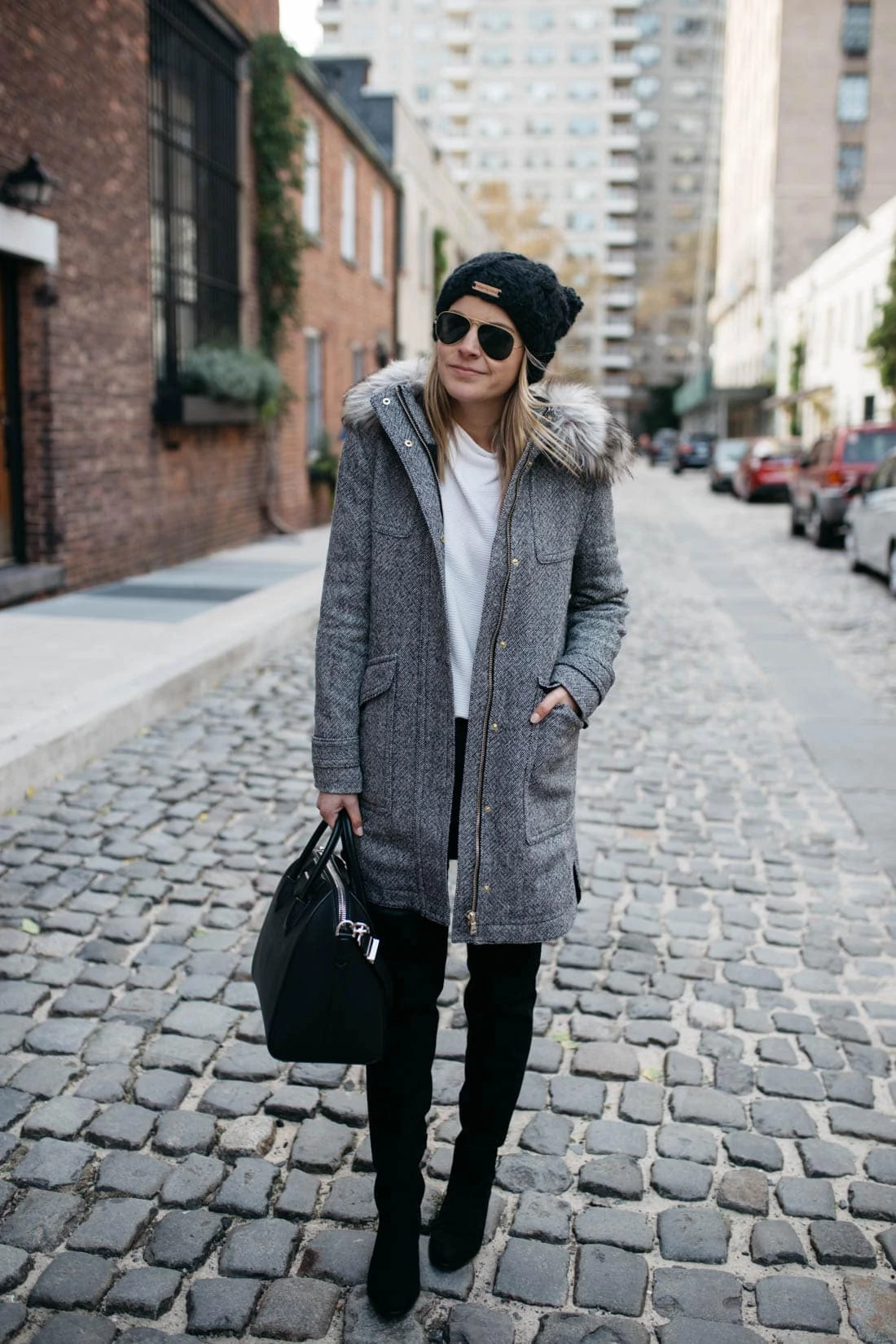 Woman in a gray tweed fur-hood parka with over-the-knee boots and a black handbag