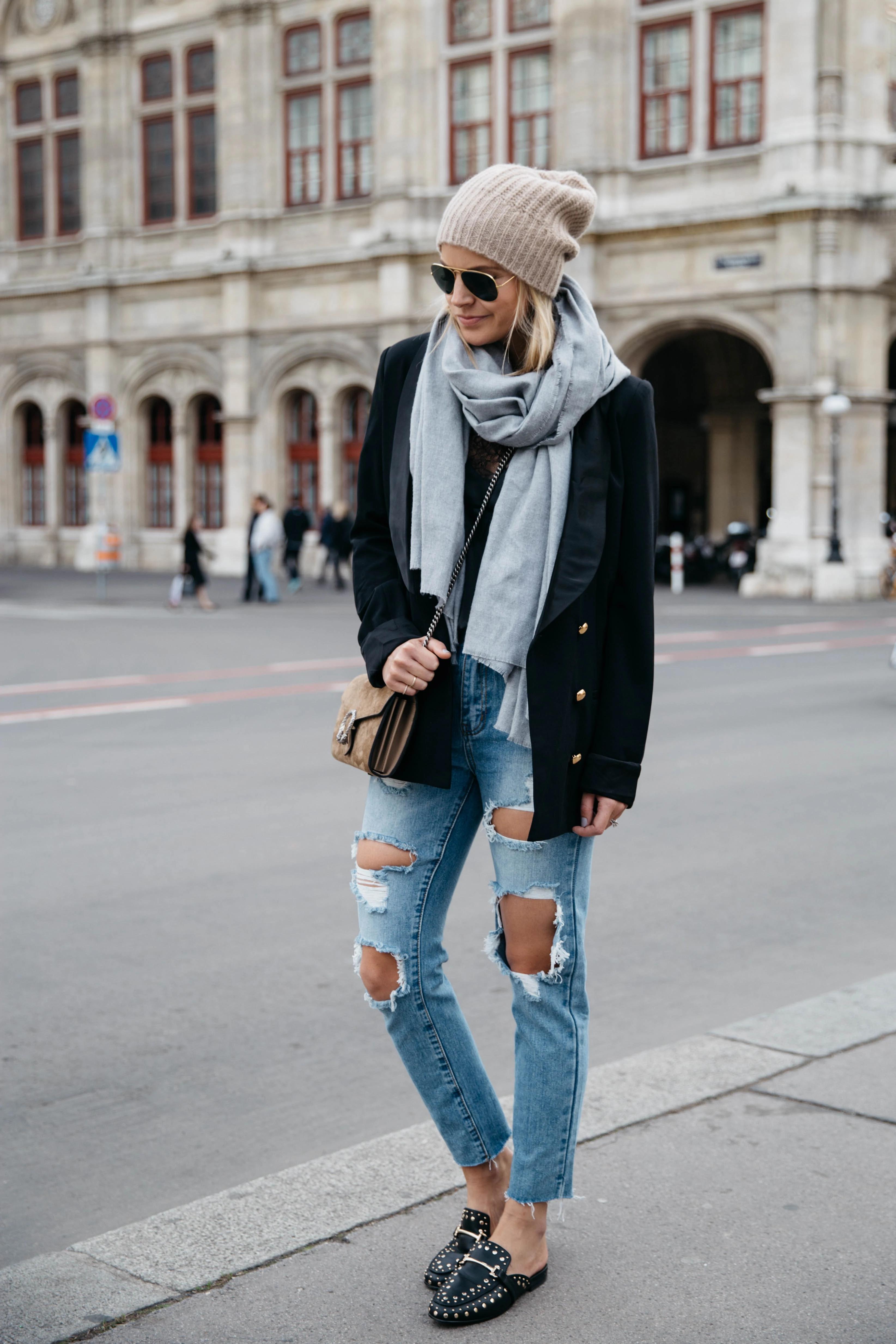 Woman in an oversized black blazer, grey scarf, knit beanie and ripped jeans with loafers