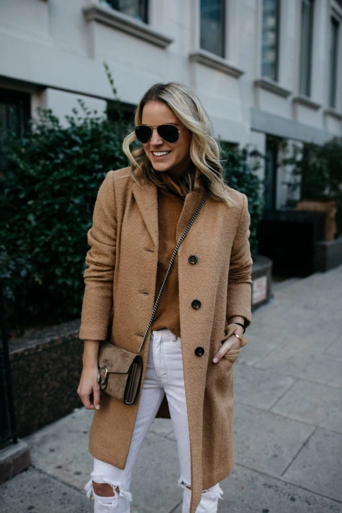 Woman wearing a camel coat over a camel turtleneck with white ripped jeans and a tan crossbody bag