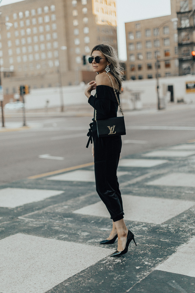 Woman in black velvet joggers with a black off-shoulder top, heels and a designer bag