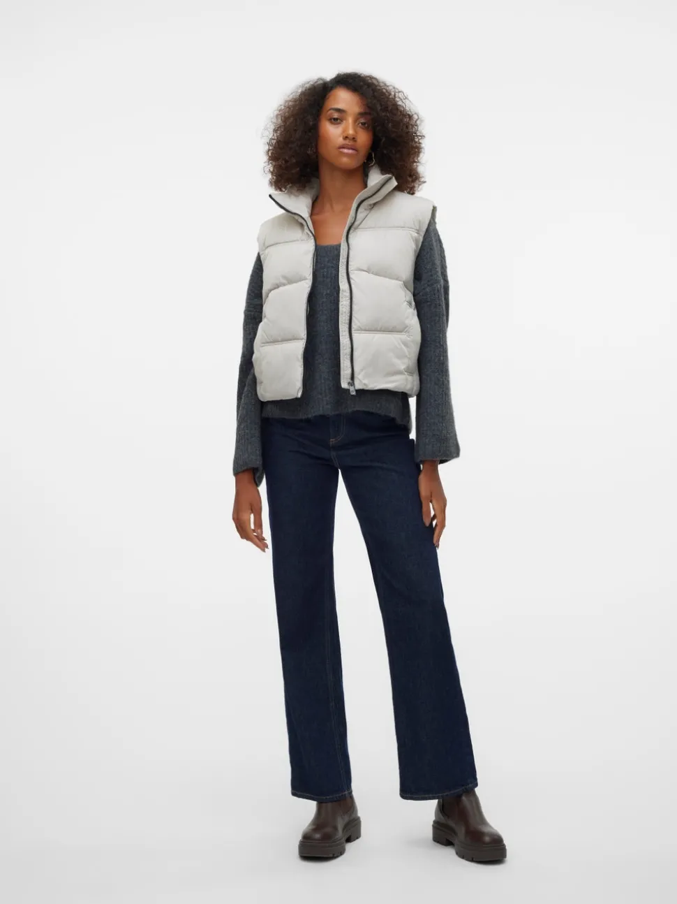 Woman in a light grey puffer vest over a grey knit with dark flared jeans and brown boots