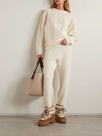 Woman wearing a cream cable-knit cropped sweater and matching knit pants, tan lace-up hiking boots and a woven tote bag