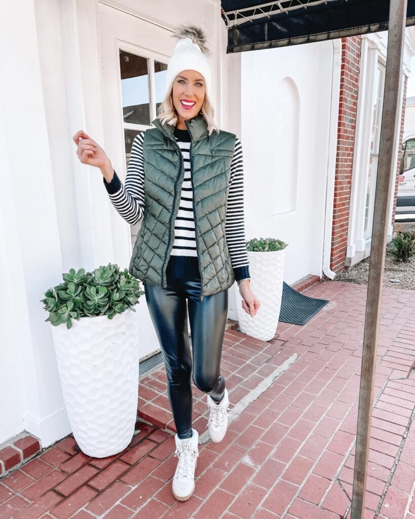 Woman in a black-and-white striped long-sleeve top, olive quilted vest, black leather pants, and white sneakers