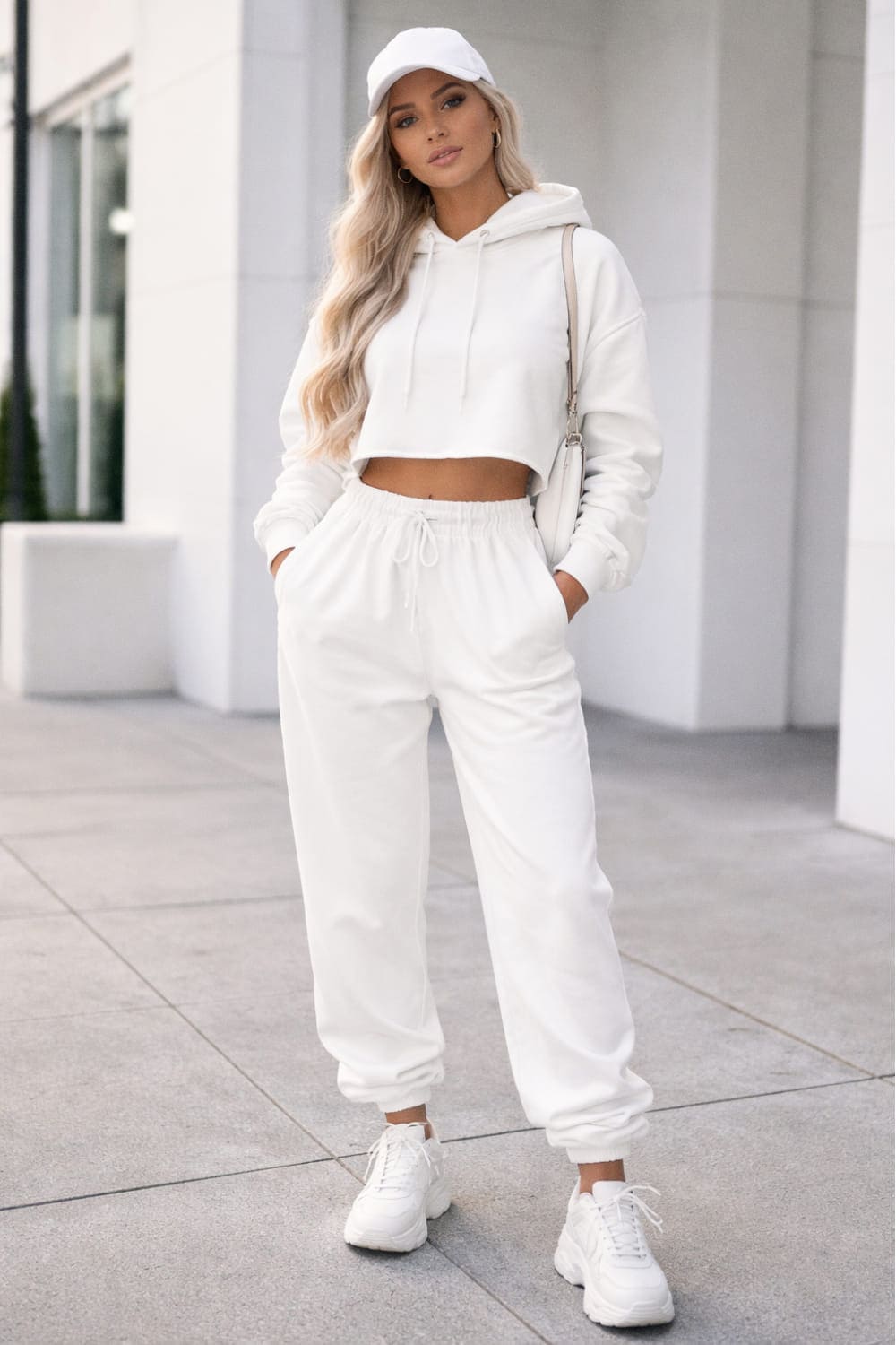Woman wearing a white cropped hoodie, white drawstring sweatpants, white sneakers, a white cap and a crossbody bag