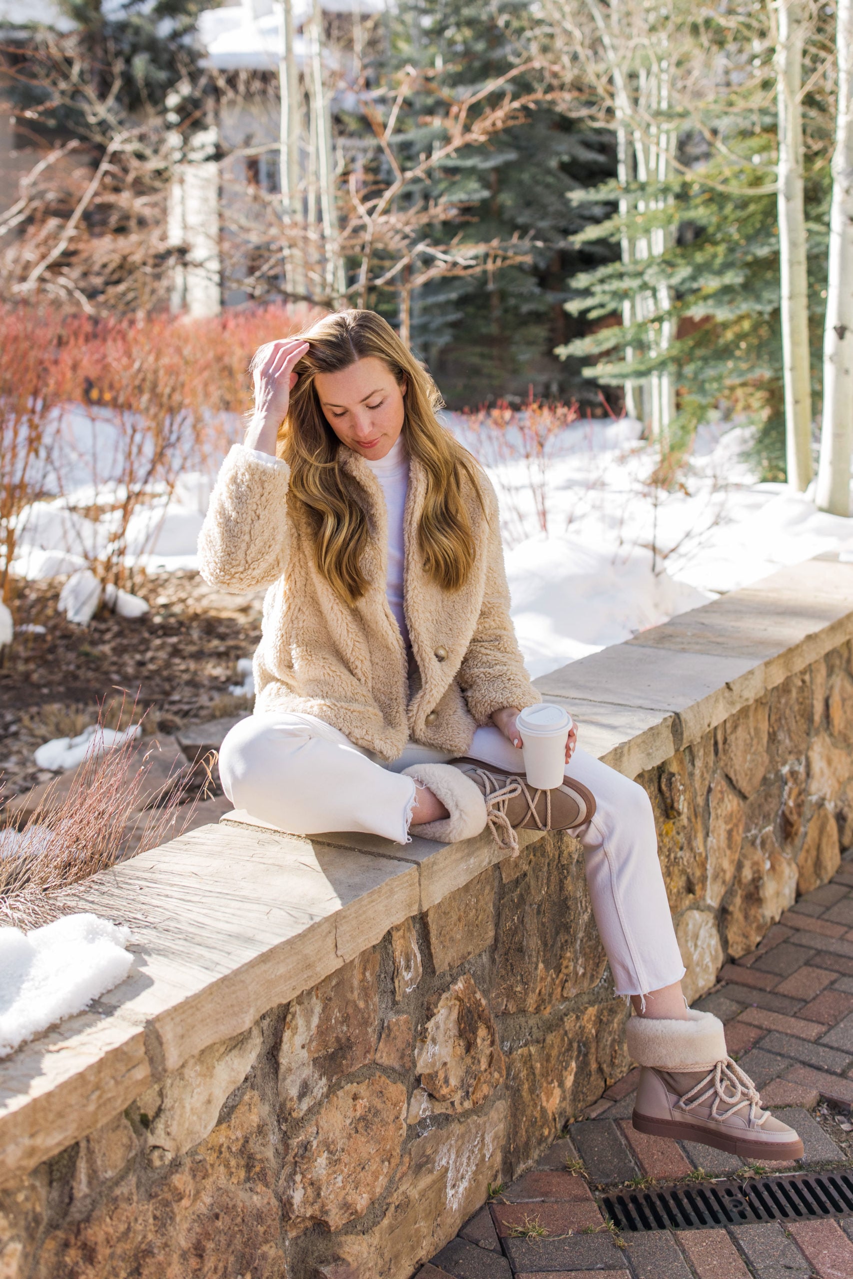 Woman in a cream fleece jacket, white turtleneck, white jeans and shearling winter boots sitting on a stone wall in the snow