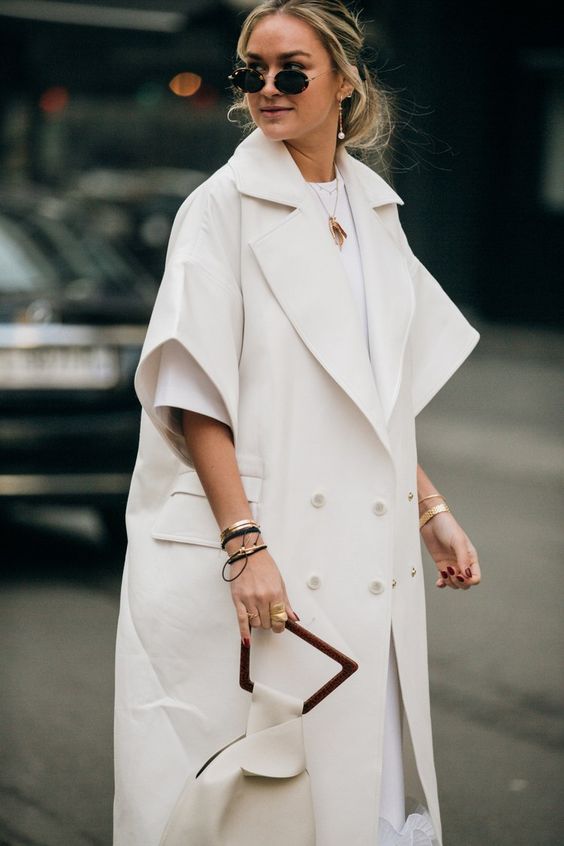 Woman in a white oversized trench vest over a white top with a white structured handbag, sunglasses and gold jewelry