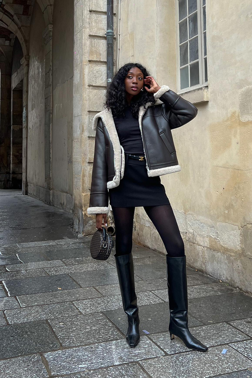 Woman in a shearling-lined black leather aviator jacket with a black mini skirt, tights and heeled knee boots