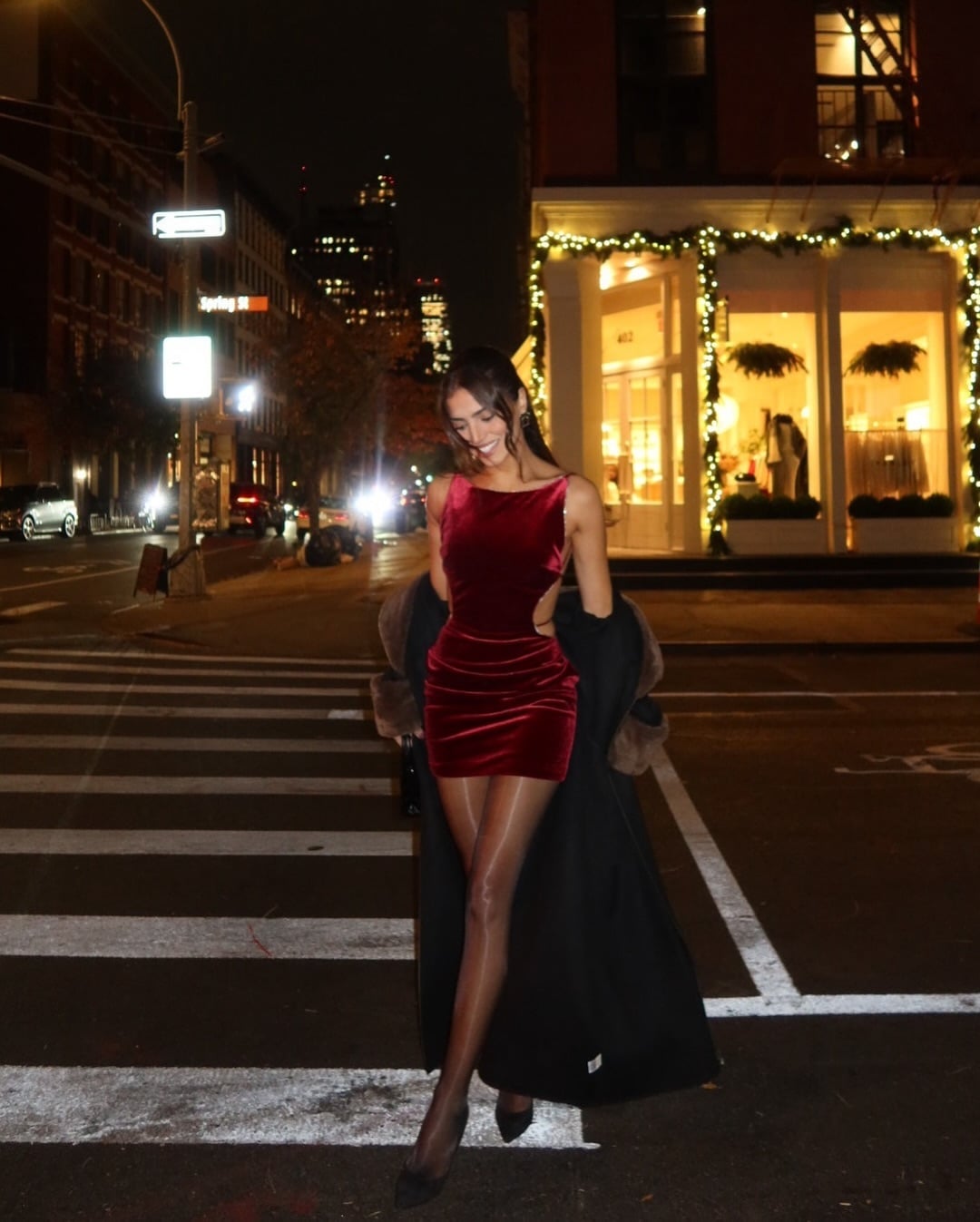 Woman in a red velvet mini dress with a long black coat and black pumps on a city street at night