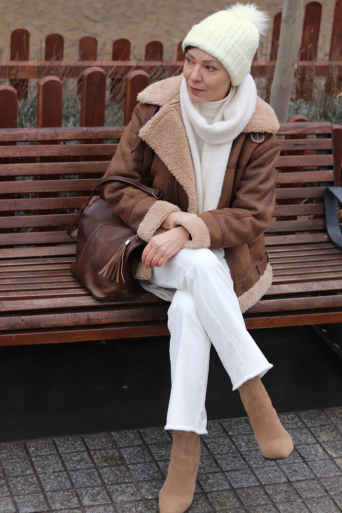 Woman in a brown shearling aviator jacket with a white knit sweater, white jeans and a knit beanie
